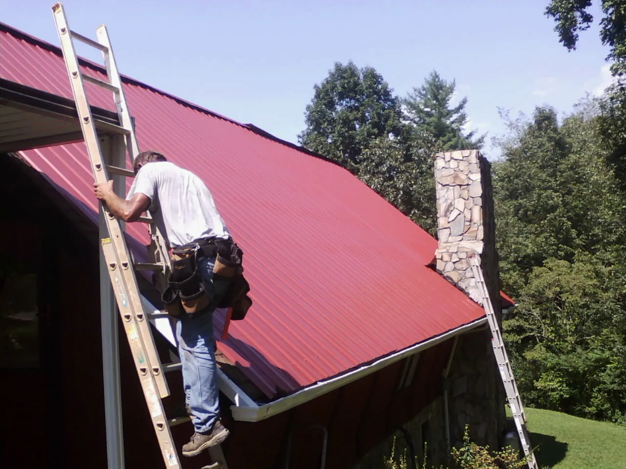 Roofer on a ladder beside a red metal roof and stone chimney — Soffit & Fascia Repair by Roofing Brothers in Highmore